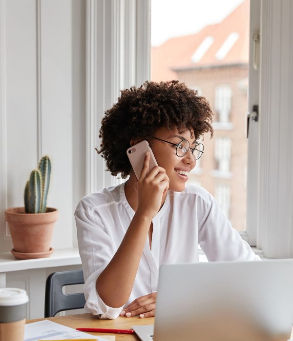 Vertical shot of cheerful woman bookkeeper talks on mobile phone, has positive expression, dressed in white stylish blouse, works with laptop computer, has work break, drinks hot aromatic beverage